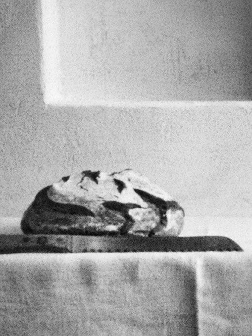 Hubertus Schülers schwarz-weiße Fotografie zeigt ein Brot, das neben einem Messer auf dem Tisch liegt.